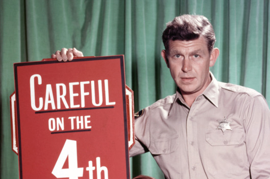 Andy Griffith, US actor, and Ron Howard, US actor, pose beside a large firework, with a sign reading 'Careful on the 4th' in a publicity portrait issued for the US television series, 'The Andy Griffith Show', USA, circa 1963. The sitcom starred