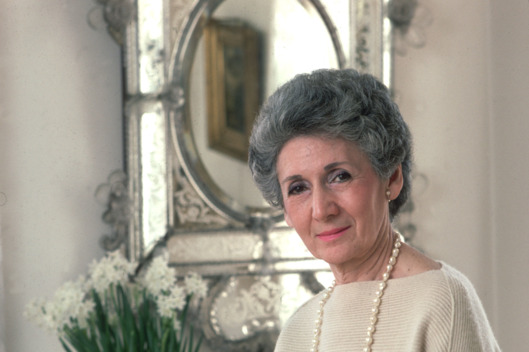 1986: A portrait of architecture critic Ada Louise Huxtable seated on a table next to a sofa with flowers and a mirror in the background.